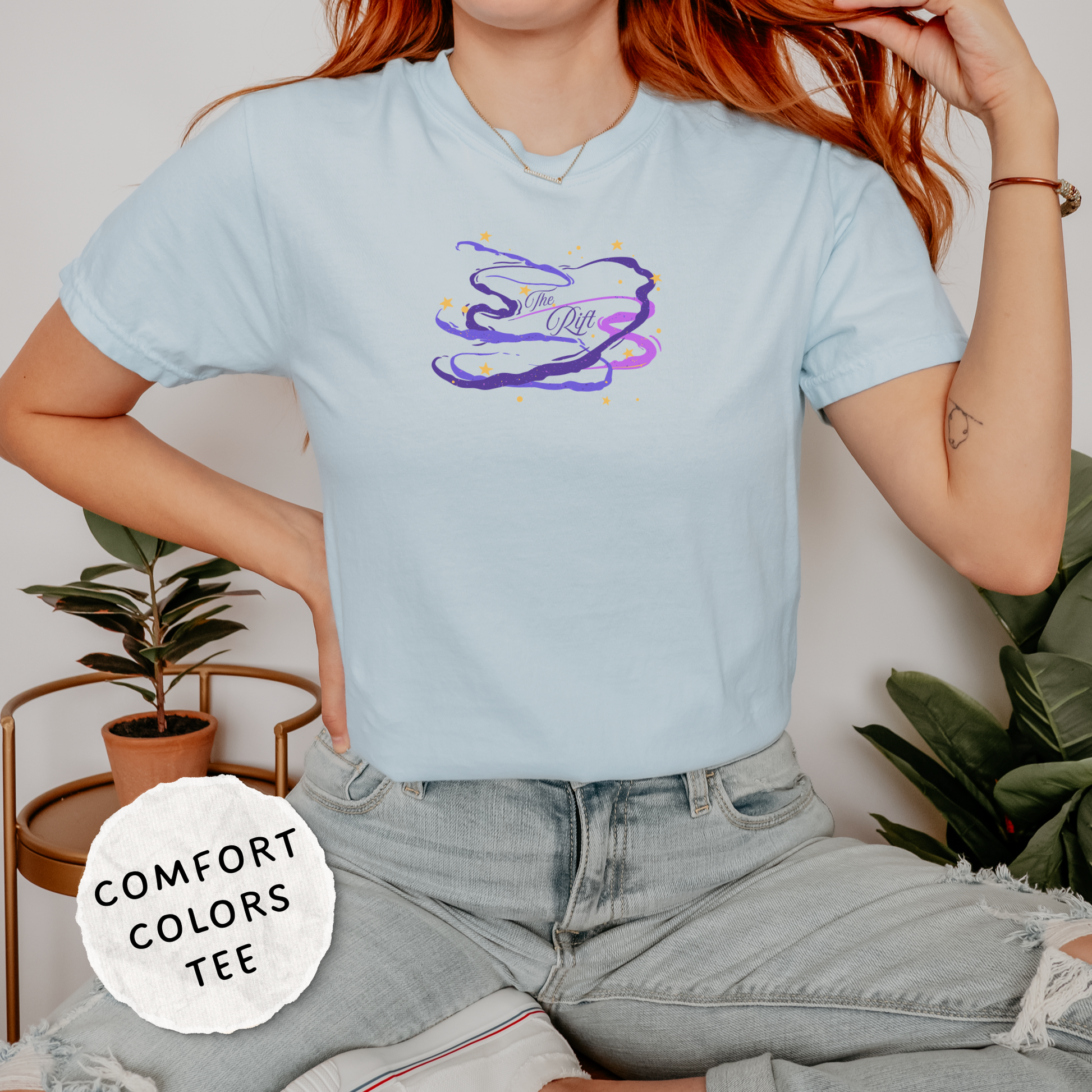 A woman wearing a light blue Comfort Colors graphic t-shirt with a purple and yellow abstract design on the chest, paired with light-wash jeans in a styled indoor setting.