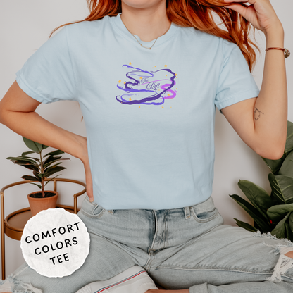 A woman wearing a light blue Comfort Colors graphic t-shirt with a purple and yellow abstract design on the chest, paired with light-wash jeans in a styled indoor setting.