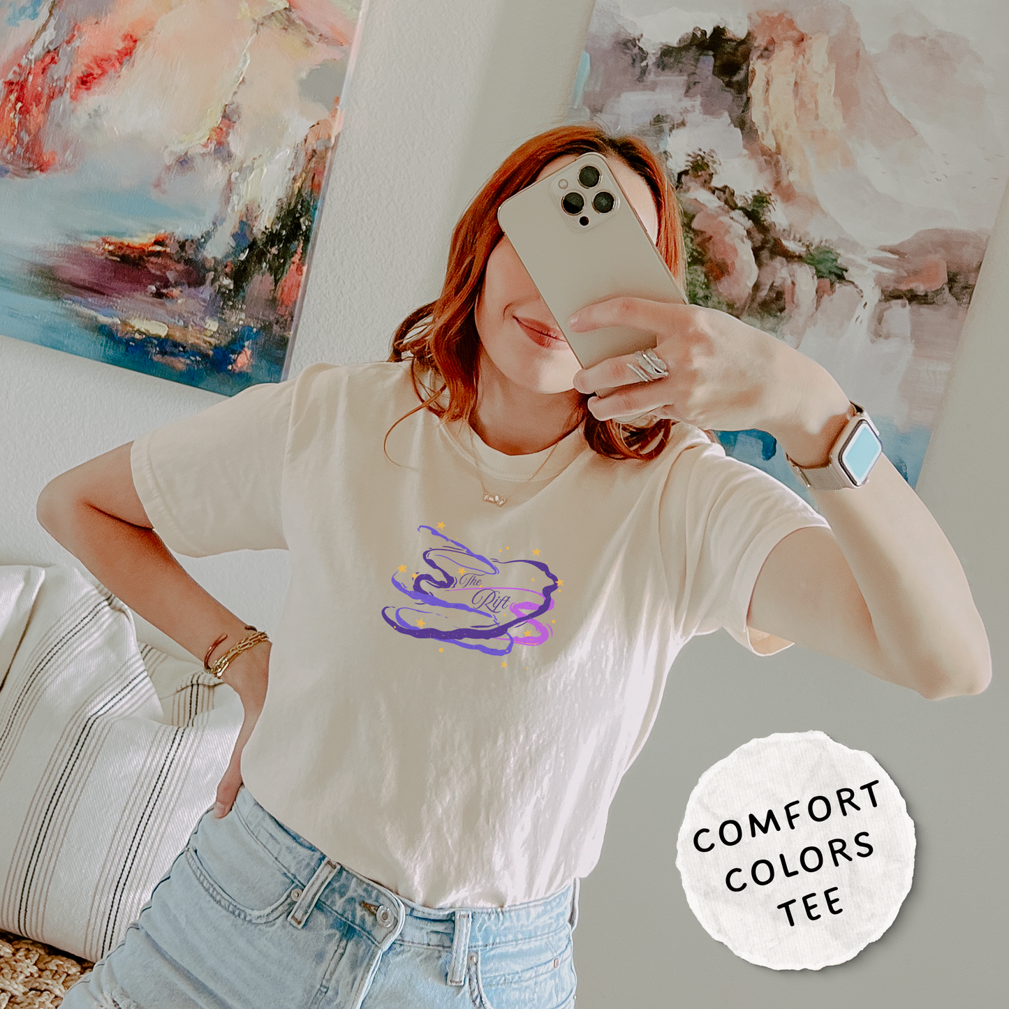 A woman takes a mirror selfie wearing a cream-colored short-sleeve t-shirt with a purple and blue abstract graphic on the front. The background shows a cozy room setting with abstract paintings on the wall and a striped pillow.
