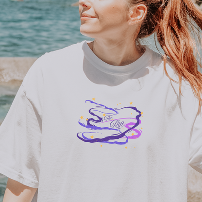 A woman wearing an oversized white short-sleeve t-shirt with a purple and blue abstract graphic on the chest, standing on a rocky beach with water in the background. A circular badge in the bottom right corner reads 'COMFORT COLORS TEE'.
