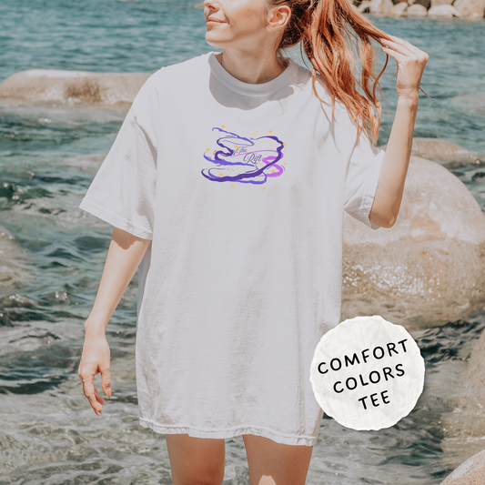 A woman wearing an oversized white short-sleeve t-shirt with a purple and blue abstract graphic on the chest, standing on a rocky beach with water in the background. A circular badge in the bottom right corner reads 'COMFORT COLORS TEE'.